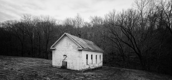 Black and white photograph of a little white abandoned building on the edge of the woods. We have no information but it looks like it may have once been a church or a school