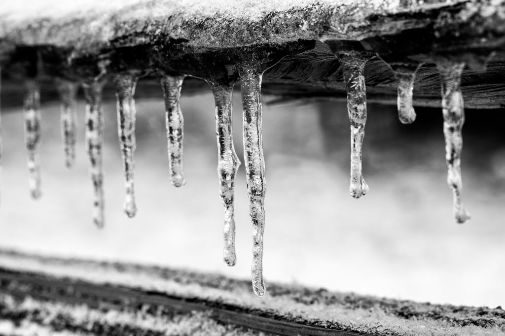 Black and white close-up photograph of a row of sparkling icicles hanging from the edge of a rustic split rail wooden fence.