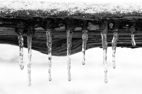Black and white macro photograph of a group of sparkling icicles hanging from the edge of a rustic split rail wooden fence.