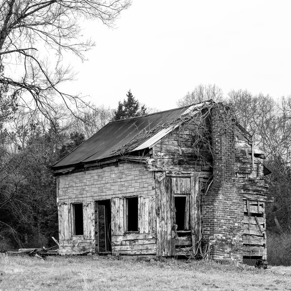 Black and white photograph of a derelict old farm house found along a stretch of isolated back road in the countryside. We can see layers of textured siding, including timbers, wooden planks, and asphalt sheets that look like bricks. (Square format)