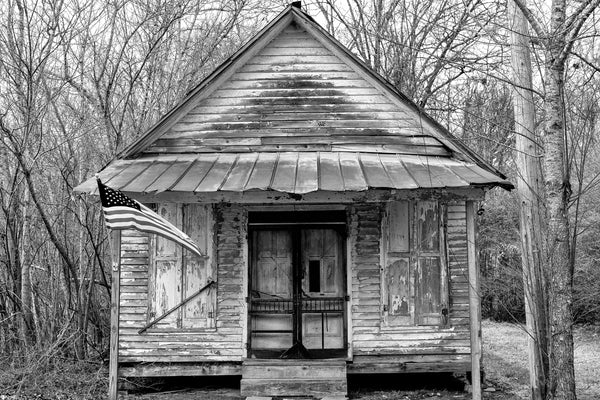 Black and white photograph of the front of an abandoned historic mercantile store found along a back road in the deep south with a rusty tin awning, peeling paint, and screen doors.