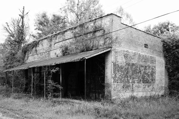 Black and white photograph of the ruined commercial storefronts with a big ghost sign on the wall in the small community of Hushpuckena, Mississippi.