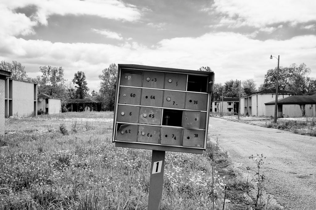Black and white photograph of broken mailboxes with an eerie, abandoned apartment complex seen in the background.