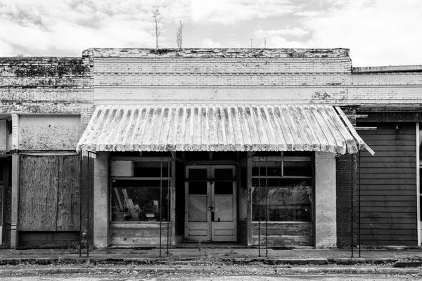 Black and white photograph of a vacant small town storefront with a rusty metal awning and plants growing from its roof.