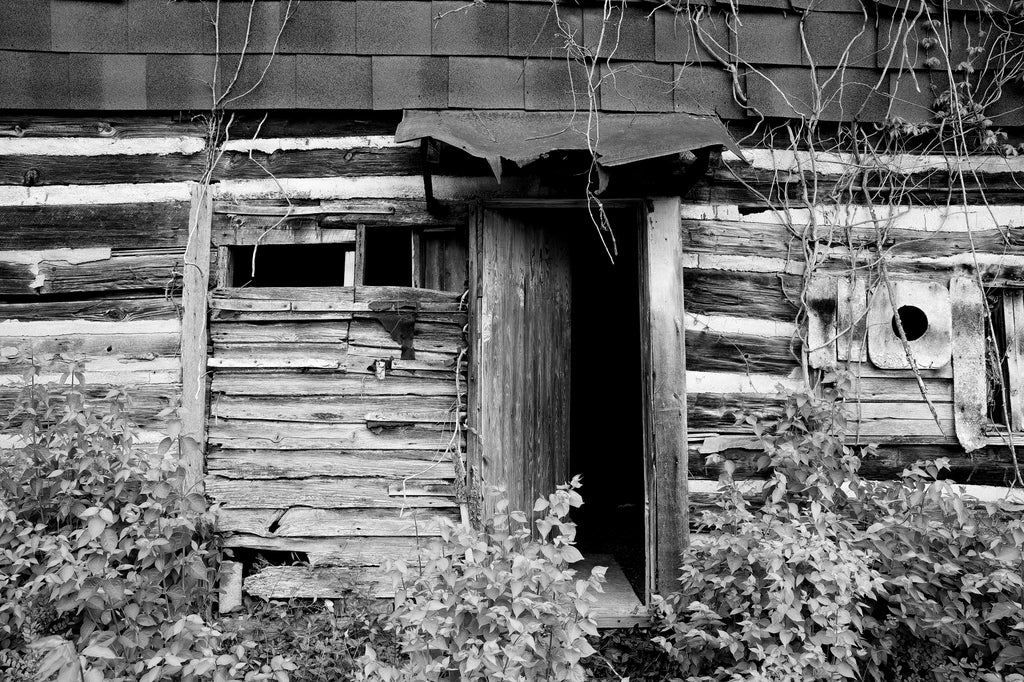 Black and white photograph of the open doorway on the ramshackle exterior of a rustic old wooden cabin that may have been a sharecropper house a long time ago