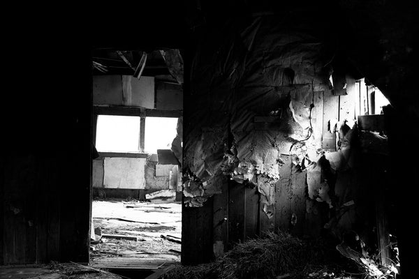 Black and white photograph of a shaft of sunlight inside a dark abandoned old house that still has hay on the floor from being used as a barn after people no longer lived there.