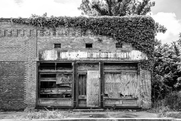 Black and white photograph of an abandoned old retail store with ivy and weathered plywood found in Clarksdale, Mississippi.
