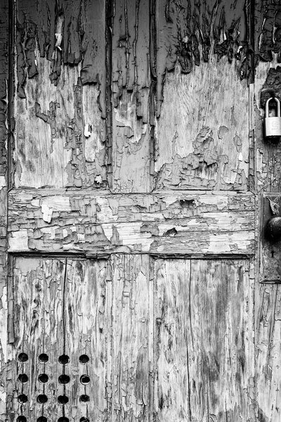 Black and white photograph of the weathered front door of an abandoned building covered in the texture of cracked and peeling paint.