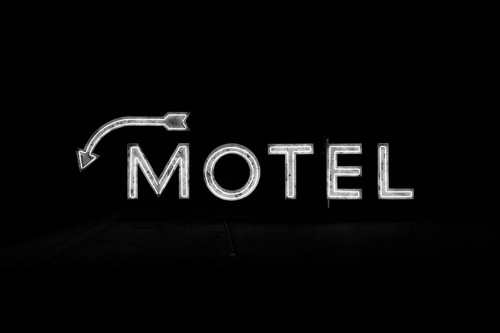 Black and white photograph of a vintage neon motel sign glowing in the dark above the roof of an old fashioned roadside motel.