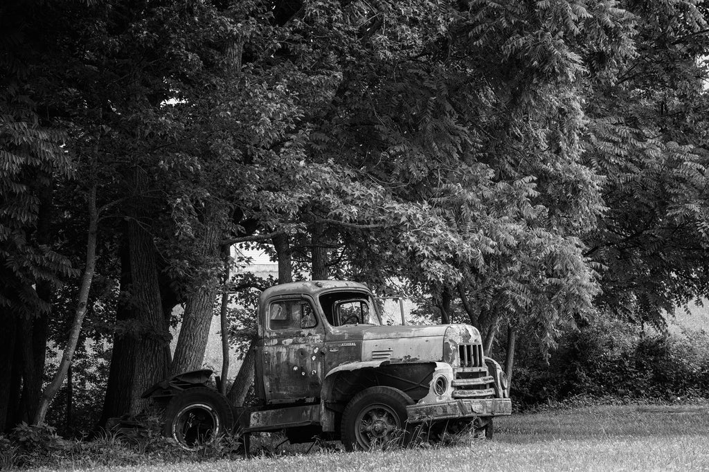Black and white photograph of a rusty old truck abandoned along a rural highway with numerous bullet holes in the metal and glass.