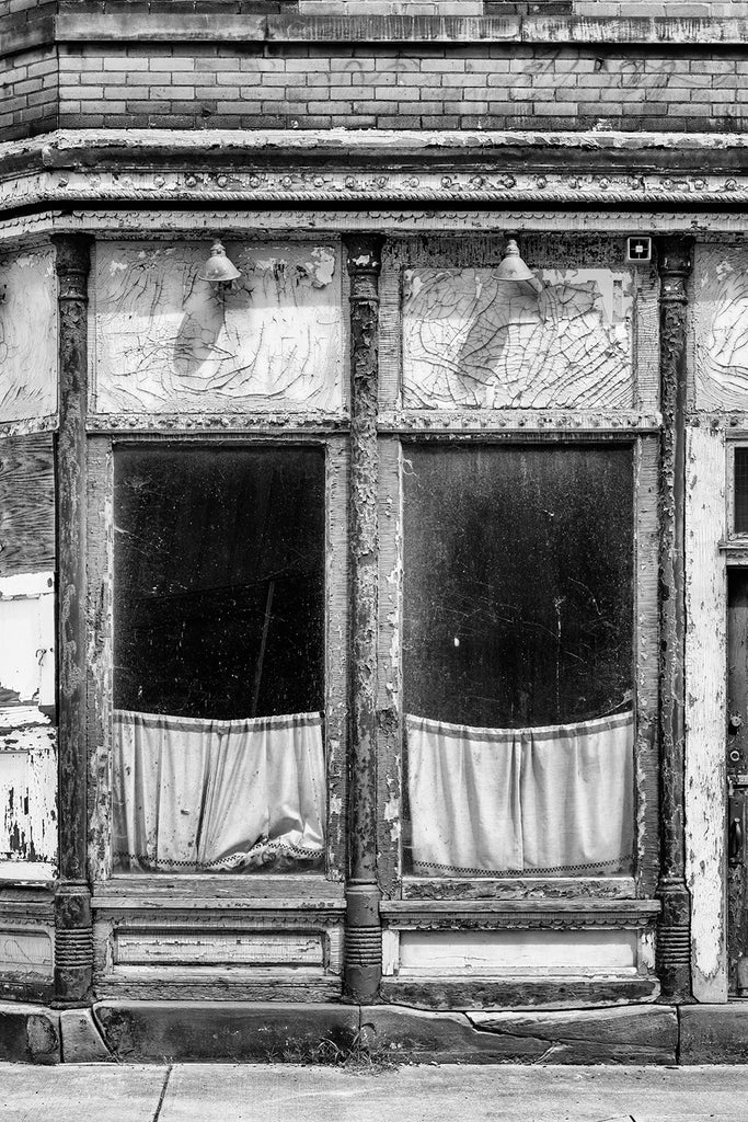 Black and white photograph of an abandoned building with cracked paint and curtains behind two dirty windows.