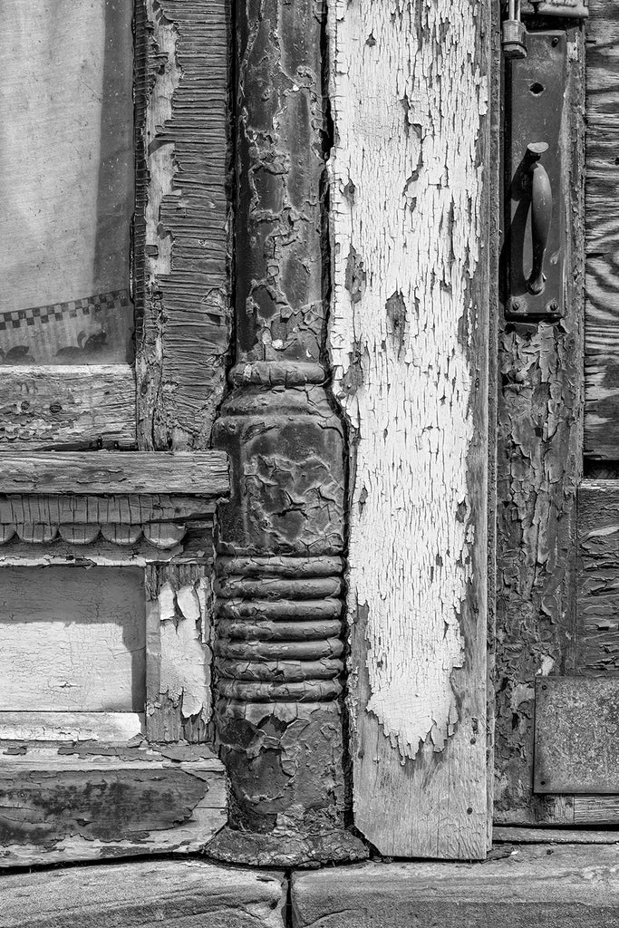 Black and white photograph of chipped paint and cracked wood on the exterior of an abandoned building that started as a merchant tailor shop owned by a German immigrant named August Helmbrecht in the 1890s.