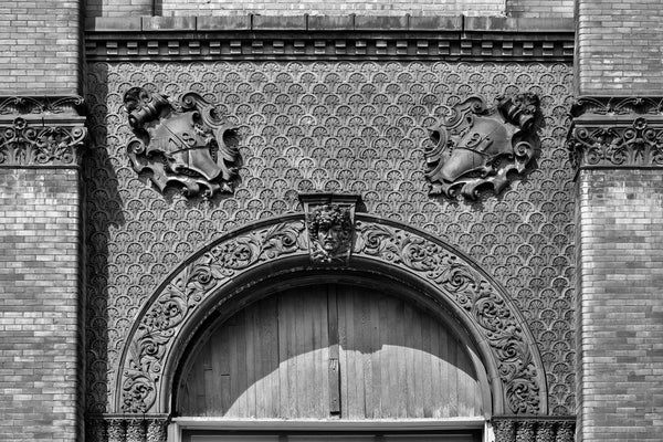 Black and white photograph of architectural details featuring a human head surrounded by ornate tile patterns on the exterior of the historic Schmulbach Brewing Company. Founded as the Nail City Brewery in 1855, it was acquired by investors in 1873. Majority partner Henry Schmulbach changed the name in 1882 and hired a master brewer from the famous Gambrinus Brewery in Cincinnati. This structure was built in 1891, and Schmulbach was closed in 1914 when West Virginia passed prohibition laws.