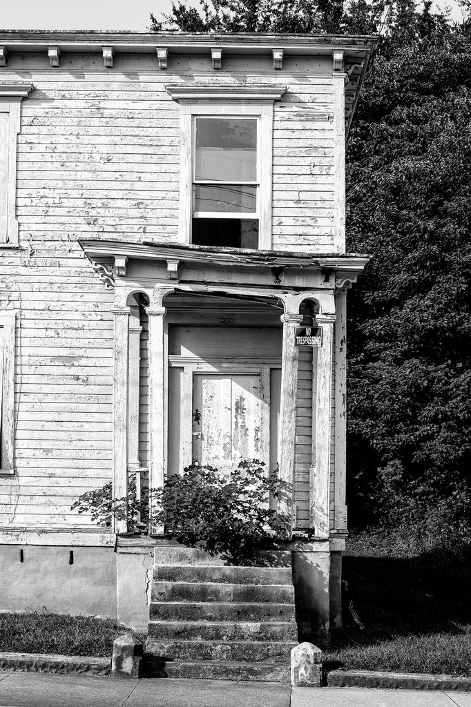 Black and white photograph of the front of an abandoned historic wooden row house with a small tree growing on its front steps, seen in Danville, Virginia.
