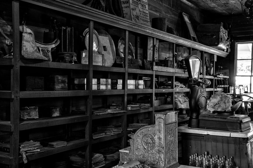 Black and white photograph of the interior of an old country store that has been preserved with merchandise on the shelves and with an antique cash register.