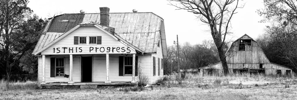 Black and white panoramic landscape photograph showing an abandoned farmhouse with a barn in the background. The house has been painted with black spray paint across its front with the question "Is this progress?"