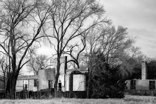 Black and white photograph of the ruins of a long-abandoned school standing among a grove of trees. The school was built by the WPA in the 1930s in the farming community of Hext, Oklahoma, and stood vacant for decades before burning in 2021.