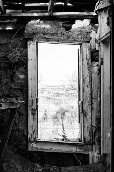 Black and white photograph of the wide open New Mexico landscape seen through the broken window frame of an abandoned motel along an isolated stretch of old Route 66