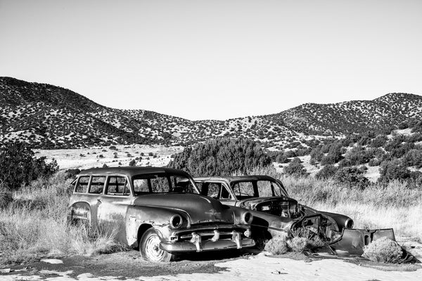 Black and white photograph of the New Mexico landscape with two abandoned rusty old automobiles and mountains rolling into the background.