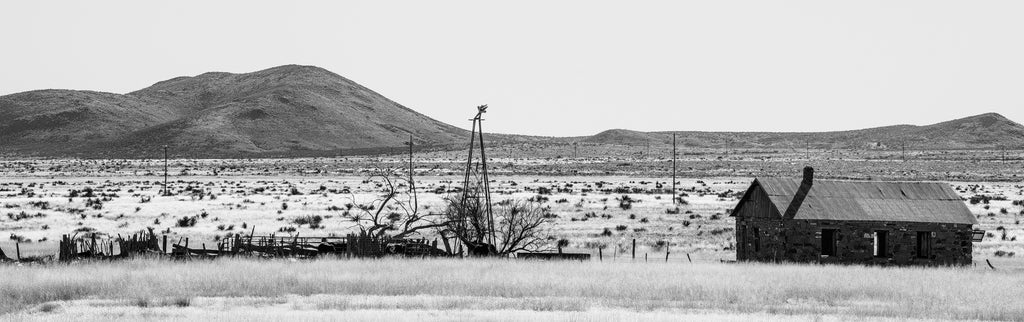 Black and white panoramic landscape photograph of an abandoned ranch house and horse corral in the vast wide high desert of New Mexico.