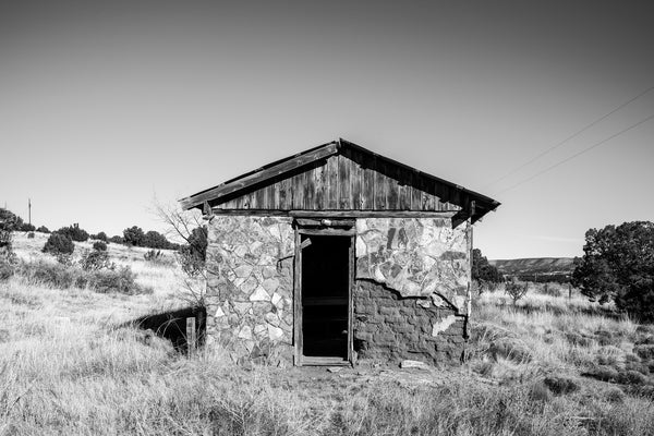 Black and white photograph of a rustic, abandoned stone and adobe house set amidst the New Mexico high desert landscape.