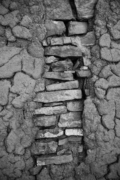 Black and white detail photograph of the textures of a weathered old adobe wall with layers of flat stones showing through. 