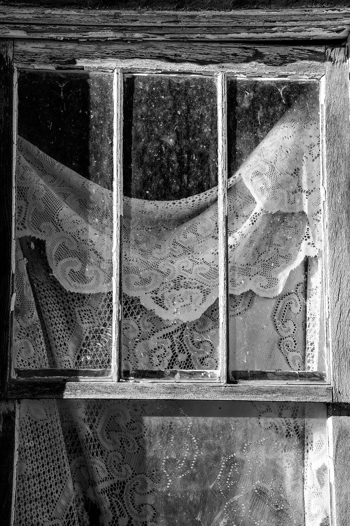 Black and white photograph of a rustic, abandoned adobe house with lace curtains in the window.