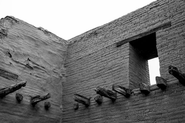 Black and white photograph of the high walls of a ruined old adobe building in the mountains of New Mexico.