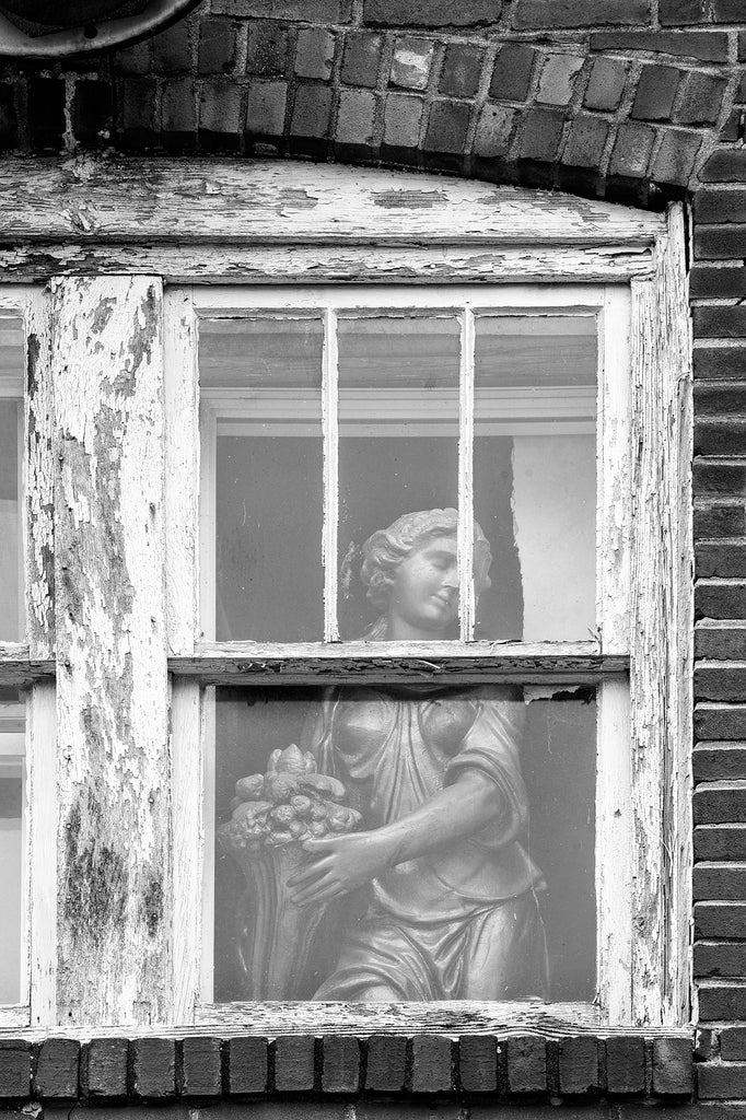 Black and white photograph of a statue of a goddess cheekily placed by someone with a sense of humor into the second-floor window of an abandoned brick building.