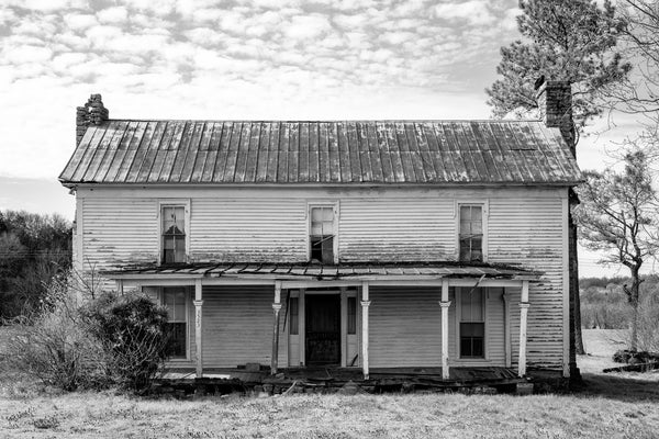 Black and white photograph of an old abandoned two-story white clapboard farmhouse with three windows across the upper floor. 