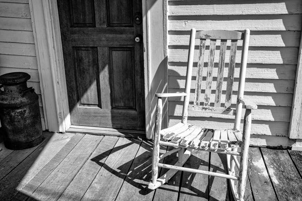 Black and white photograph of an old white wooden rocking chair sitting in the sun on a rustic front porch in the country.