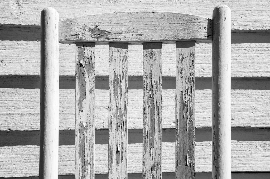 Black and white photograph of an old white wooden rocking chair juxtaposed against a white clapboard wall becomes almost an exercise in abstraction.