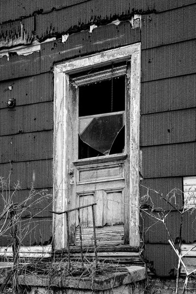 Black and white photograph of a weathered white door set amidst dark brown composition siding on the exterior of a large abandoned house in Cairo, Illinois.