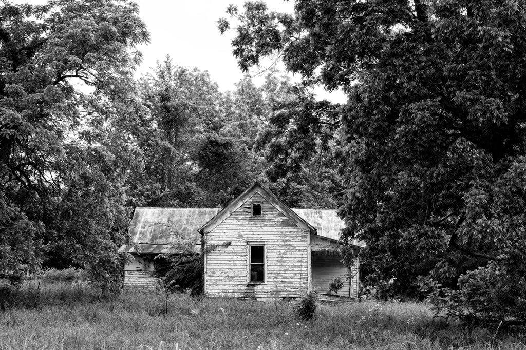 Black and white photograph of a little white abandoned farmhouse with missing windows and peeling paint.