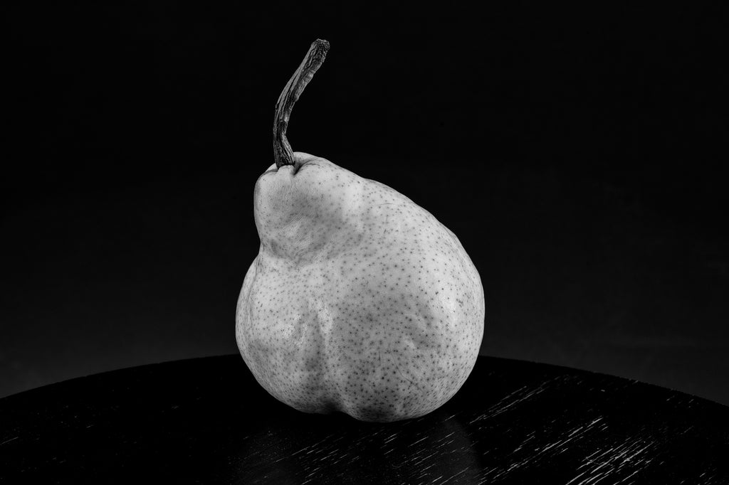 Black and white still life photograph of a plump green pear on a black table.