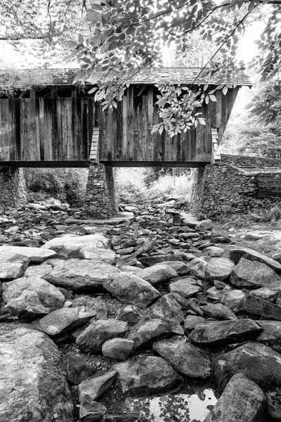 Black and white photograph of an old wooden covered bridge crossing over a rocky riverbed.