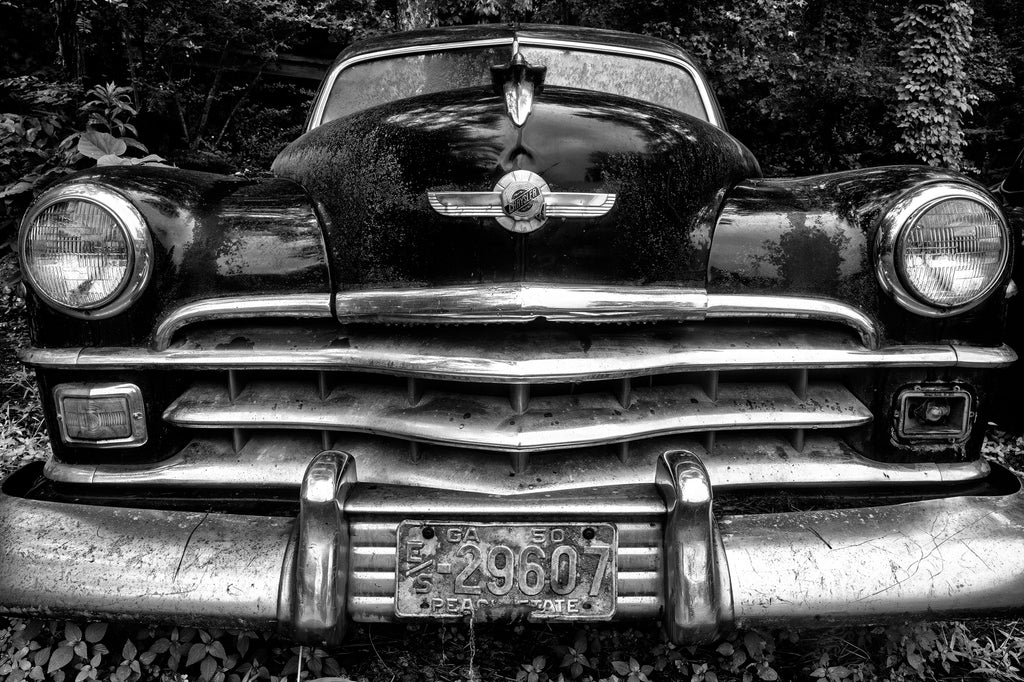 Black and white photograph of the imposing front grill and headlights of an antique black Chrysler in a junkyard with Georgia license plates dated 1950.