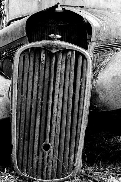 Black and white photograph of the rusty front grill of a historic Singer brand car found in a junkyard. Singer was a British company that began manufacturing bicycles in 1874 and automobiles beginning in 1901.