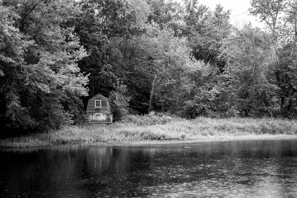 Black and white photograph of a gentle rain on the Concord River with a historic boathouse in the background.