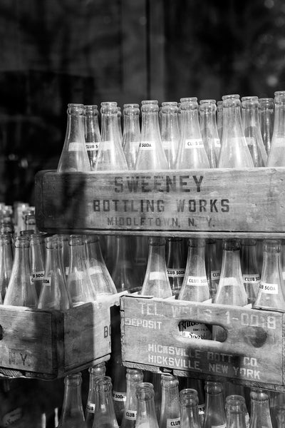 Black and white photograph of crates filled with dusty club soda bottles stacked behind the window of a vacant storefront in a small town.