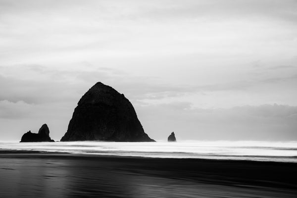 Black and white photograph of the iconic Haystack Rock at Cannon Beach, Oregon. Shot on a cloudy morning just before sunrise with a 6-stop ND filter, the long exposure caused a gauzy motion blur in the rising tide.