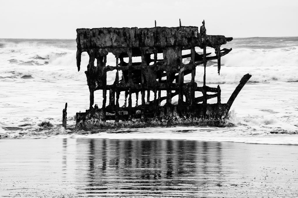Black and white photograph of the rusty bones of the wreck of the Peter Iredale on the stormy coast of Oregon near Astoria. The metal ship ran aground during strong winds in 1906 with no injuries.