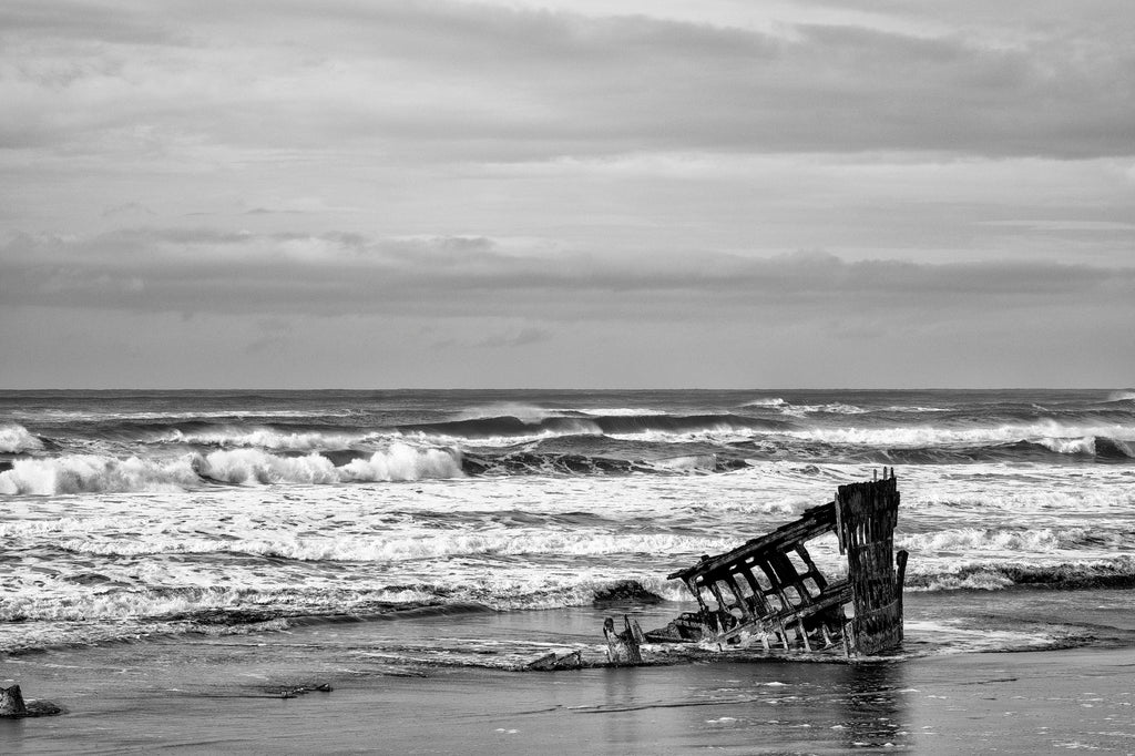 Black and white photograph of the rusty ruins of the Peter Iredale shipwreck on the stormy coast of Oregon near Astoria. The metal ship ran aground in bad weather in 1906 with no injuries.