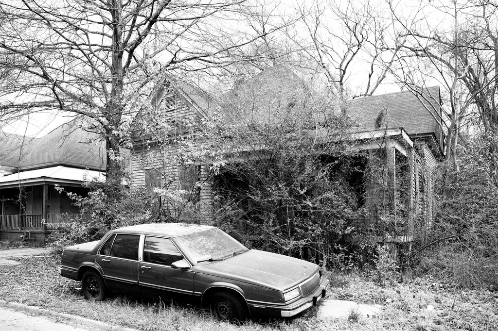 Black and white photograph of a junked car in the front yard of an abandoned house in a neighborhood of mostly derelict houses in Meridian, Mississippi.