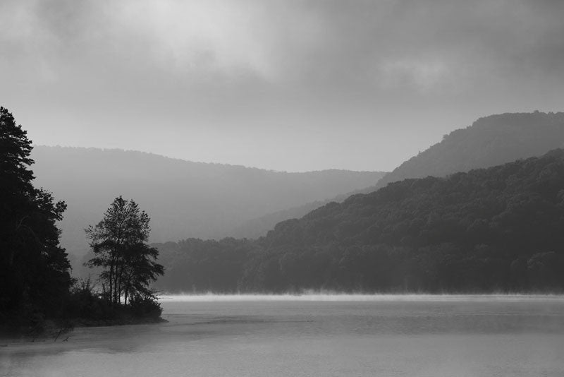 Black and white photograph of a lake surrounded by hills in early morning, as mists rise from the surface of the water, creating a mysterious mood.