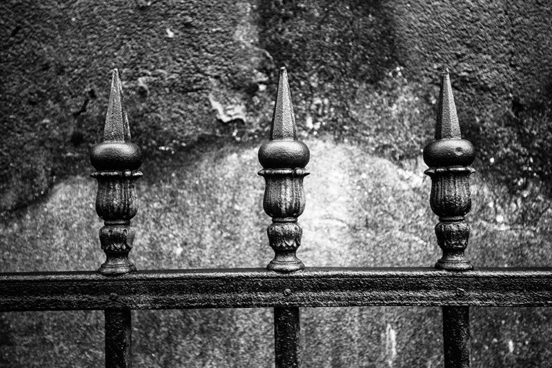 Black and white fine art photograph of three pointed pickets of an iron fence near a textural cement wall photographed on a rainy morning in New Orleans, Louisiana.