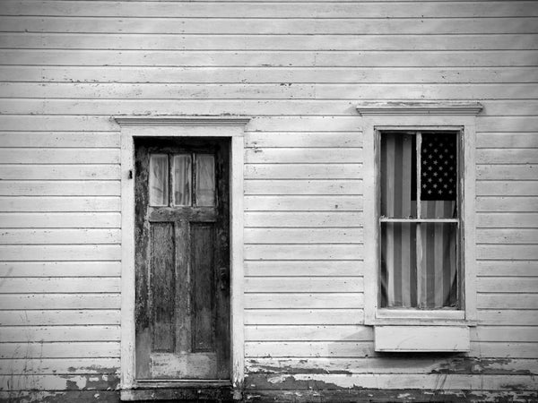 Black and white photograph of a little white house with a US flag hanging in the front window.
