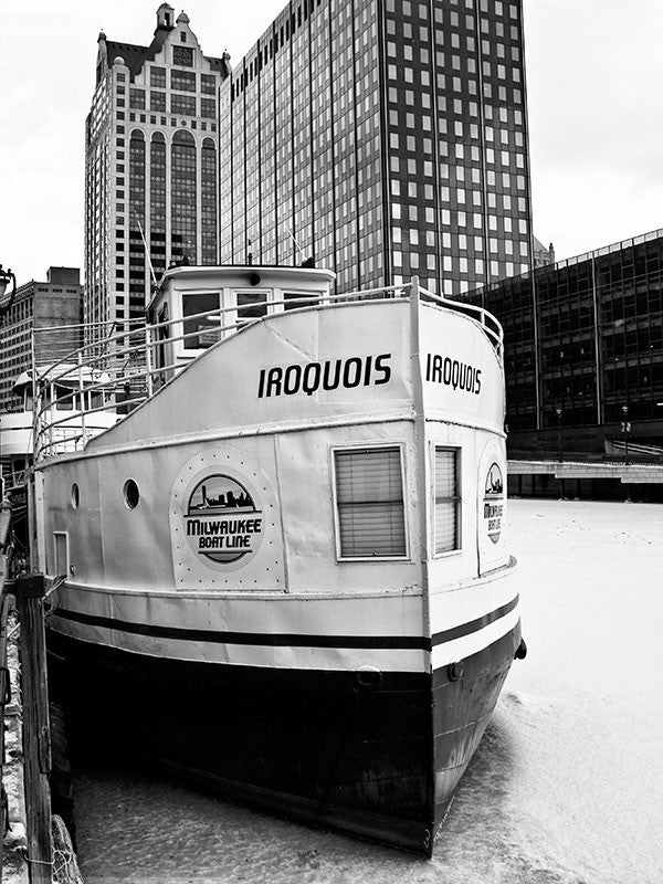 Black and white photograph of a boat named "Iroquois" frozen into the Milwaukee River on a cold winter day in downtown Milwaukee.