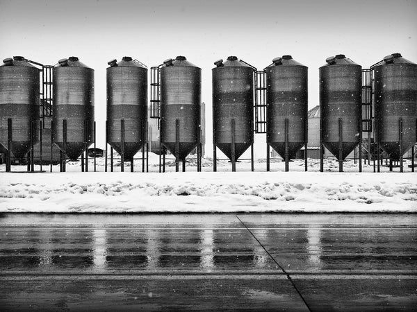 Black and white landscape photograph of a row of dark grain bins reflecting on wet pavement during an active snow storm.