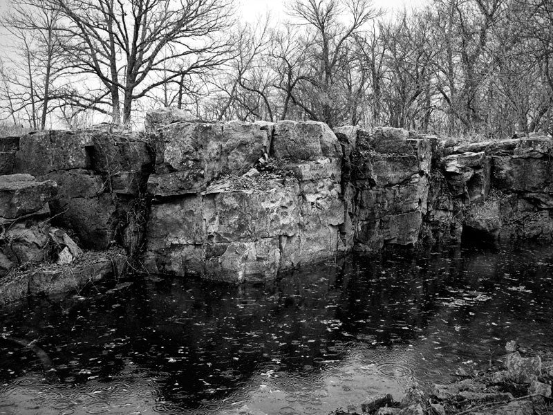 Black and white landscape photograph of the red stone quarry pit at Pipestone, Minnesota, filled with rainwater and winter meltwater. The quarry at Pipestone has been used by Native Americans for 3,000 years to mine stone to make ceremonial tobacco pipes. The site is still used for this purpose today, and is considered sacred by the tribes who use it. 